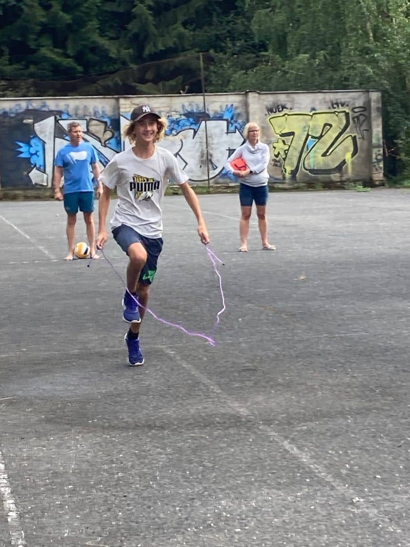 Chlapec skáče přes švihadlo, v pozadí dva dospělí a zeď s graffiti