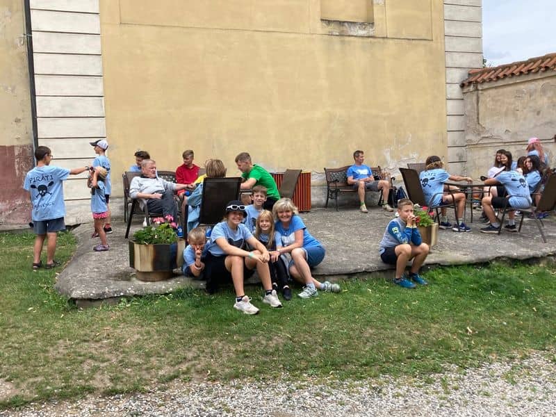 Skupina dětí a dospělých sedí venku u staré budovy