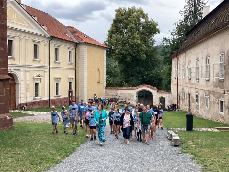 Skupina lidí prochází mezi historickými budovami po štěrkové cestě